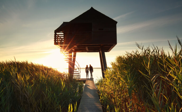 Twee mensen staan op een promenade bij een paalwoning omringd door hoog riet en kijken naar de zonsondergang over het water. Zonlicht schijnt door het bouwsel en creëert een warme, vredige sfeer.